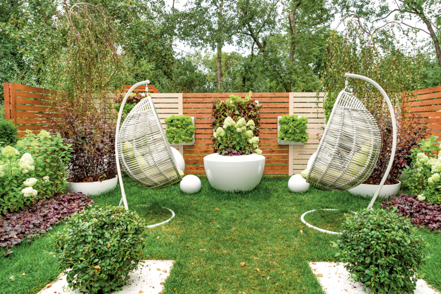 Zwei weiße, moderne Hängesessel aus Rattangeflecht am Rasen im Garten, dahinter ein moderner Holzzaun als Sichtschutz.