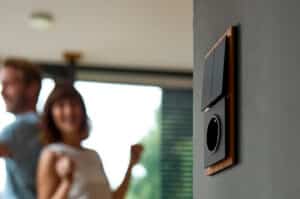 ABB zeigt einen Busch-Jaeger Doppel-Schalter mit schwarzen Wippen mit einer schwarzen Steckdose und Holz-Rahmen an einer dunkelgrauen Wand.