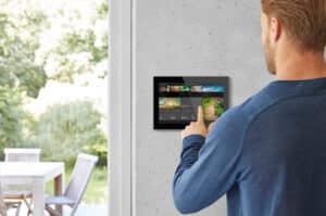 ABB zeigt eine Busch-Jaeger Steuereinheit mit großem Touch-Display in schwarz an einer grauen Wand in einem Wohnraum.