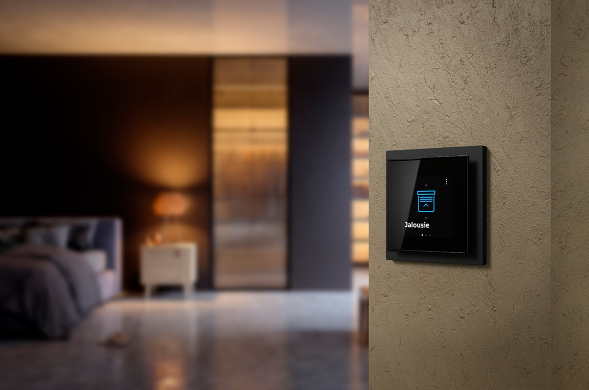 ABB zeigt einen Busch-Jaeger Regler für Jalousie in schwarz in einem Schlafzimmer.