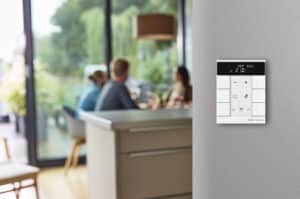 ABB zeigt eine Busch-Jaeger Steuereinheit für Temperatur mit Display in weiß an einer grauen Wand in einer Küche mit Essbereich.