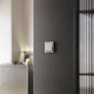 ABB zeigt einen Busch-Jaeger Schalter mit Wippe und Rahmen aus Edelstahl an einer dunkelgrauen Wand.