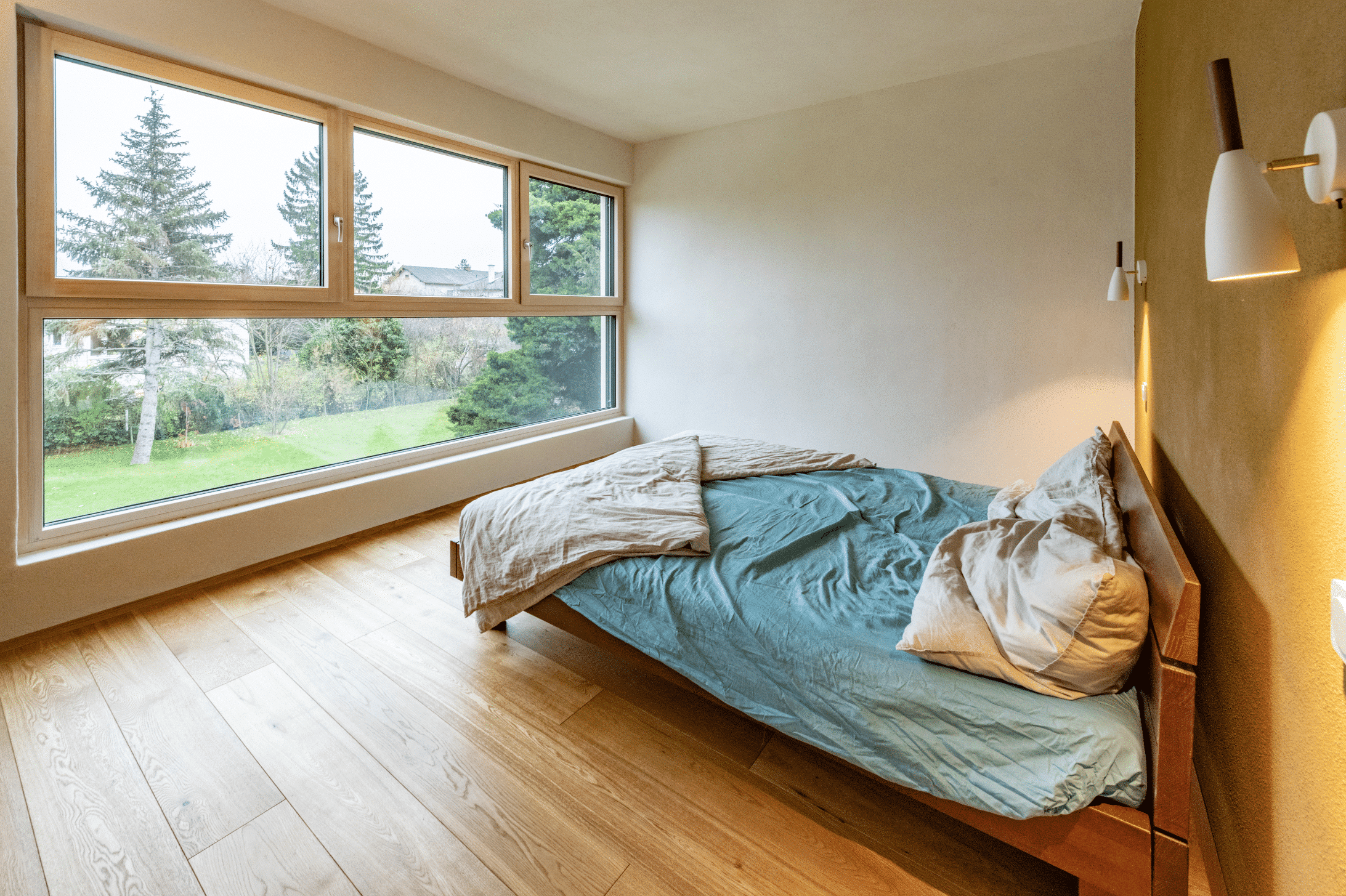 AL Architekt ZT zeigt ein kleines Schlafzimmer mit einem Doppelbett aus Holz, weissen Wandleuchten und einem großem Panorama-Fenster mit Ausblick ins Grüne.