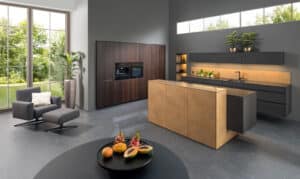 Eine grifﬂose Küchenzeile mit schwarzer Oberfläche, einer Gerätezeile aus Walnussholz und einer Kochinsel mit Theke in Bronze und bronze‑farbenem Lichtakzent von ALPHAOMEGA.