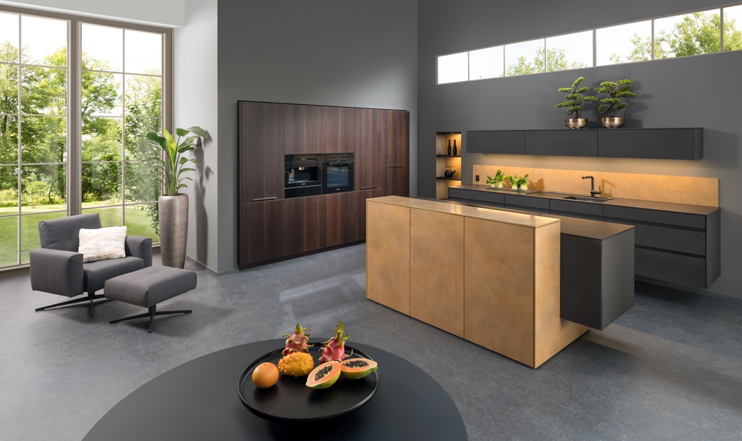 Eine grifﬂose Küchenzeile mit schwarzer Oberfläche, einer Gerätezeile aus Walnussholz und einer Kochinsel mit Theke in Bronze und bronze‑farbenem Lichtakzent von ALPHAOMEGA.