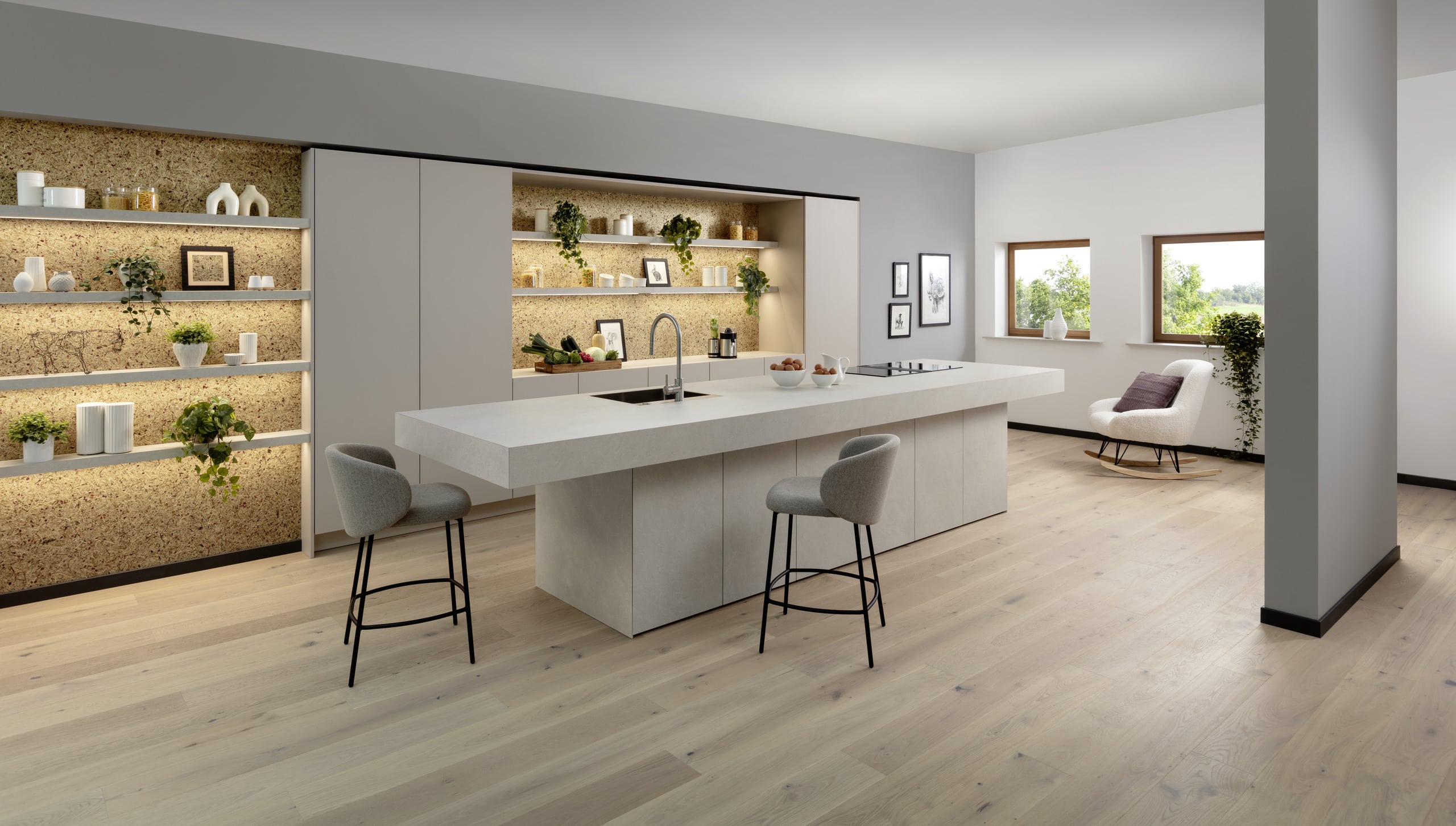 Eine grifflos gestaltete Küche mit Kochinsel in Betonoptik und einer eingekofferten Küchenzeile mit Korkrückwand, indirekter Beleuchtung und schwebenden Regalablagen von ALPHAOMEGA.