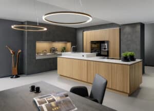 Eine grifflos gestaltete Küche mit Kochinsel und eingekofferte Küchenzeile mit Front und Rückwänden aus einer warmen geriffelten Holzfurnier und einem angrenzenden Essbereich mit großen ringförmigen Pendelleuchten von ALPHAOMEGA.