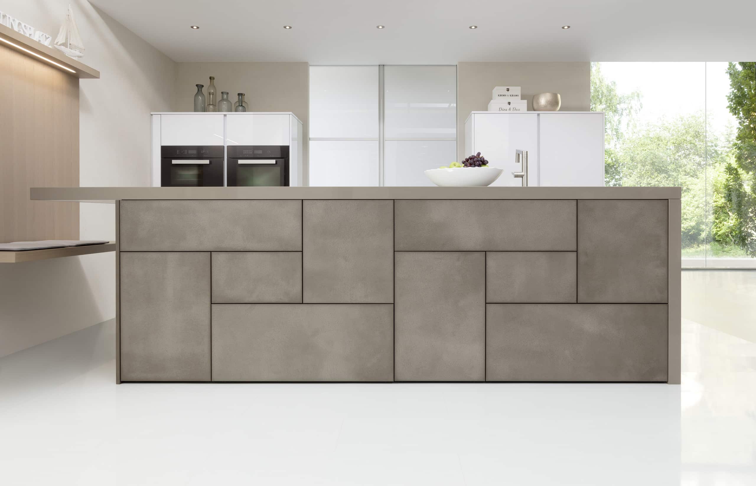 Ein griffloses, geometrisches Frontendesign an einer Kochinsel in beiger Betonoptik mit wandmontierter schwebender Sitzbank aus Holz von ALPHAOMEGA.