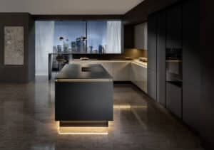 Eine grifflos gestaltete Küche mit schwarzem beleuchteten Sockel, Kochinsel mit Bar‑Theke und indirekter Beleuchtung unter der Arbeitsplatte von ALPHAOMEGA.
