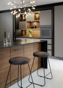 Eine Küchenzeile und Kochinsel in Grau mit schwarzer Arbeitsfläche, Inselsockel und Innenleben der Geräte­zeile aus Nussbaumholz von ALPHAOMEGA.