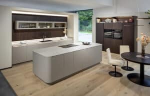 Eine grifﬂose Küchenzeile mit abgerundeter grauer Kochinsel, Küchenzeile mit Walnussrückwand, einer passenden Gerätezeile und einem runden Kaffeetisch mit Polsterdrehstühlen von ALPHAOMEGA.