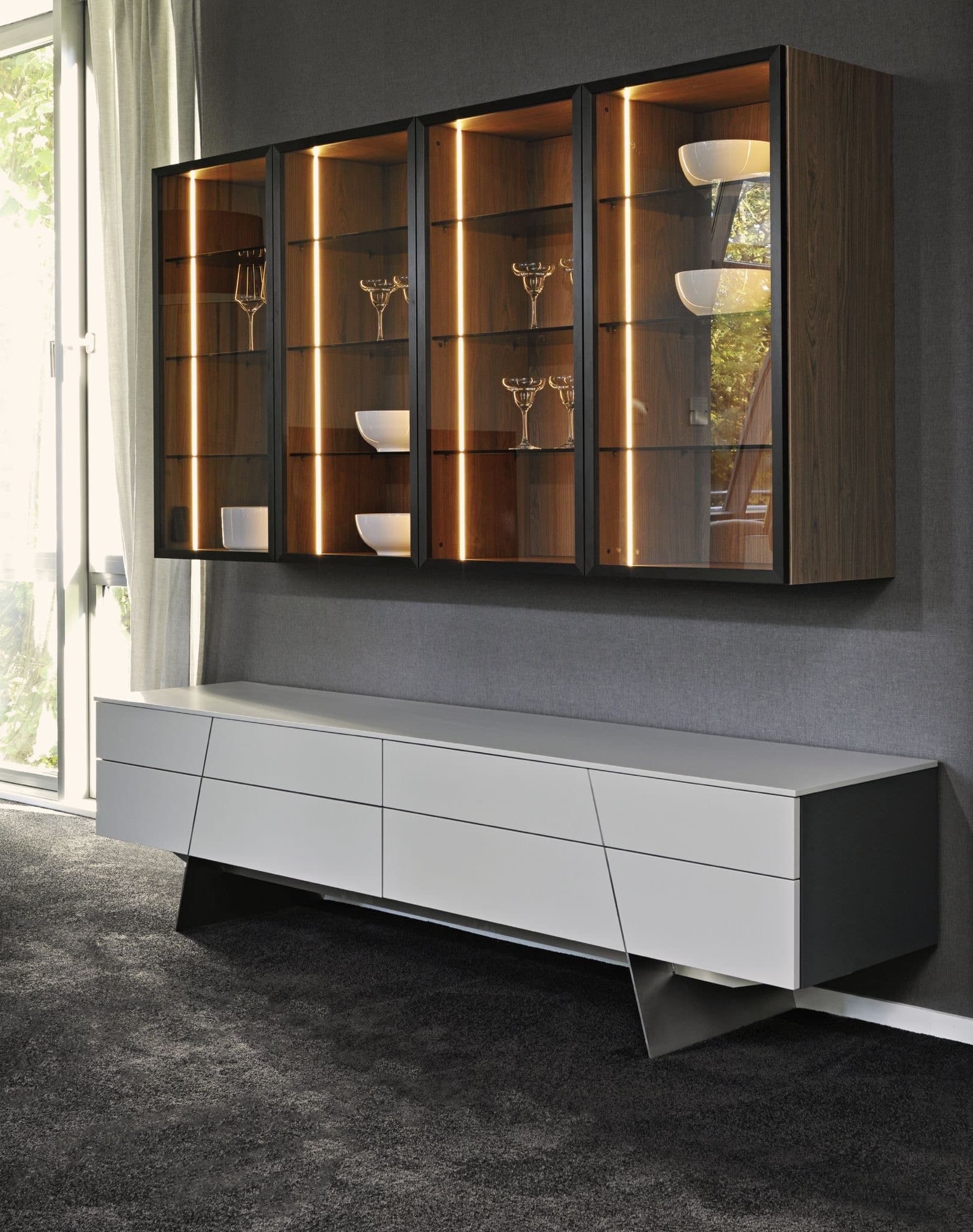 Eine Wohnzimmervitrine in warmem Holzton, Glastüren und sanftem Amberlicht, darunter ein modernes Sideboard mit silbernen Standbeinen von ALPHAOMEGA.