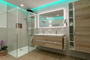 Badezimmer mit großformatigen hellgrauen Fliesen, Duschkabine mit Glasschiebetür, Regendusche und Handbrausearmatur, schwebendem Doppelwaschtisch in Holzoptik, hinterleuchtetem großformatigen Spiegel, ausziehbarem Vergrößerungsspiegel, Laminatboden und herabgesetzter Decke mit Spots und LED-Licht.
