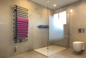 Badezimmer mit großformatigen hellgrauen Fliesen, Duschnische mit Duschtasse, schwebender Duschablage, Glaswand mit Teilsatinierung als Sichtschutz und einem wandmontiertem schwarzem Handtuchwärmer in Heizstaboptik.