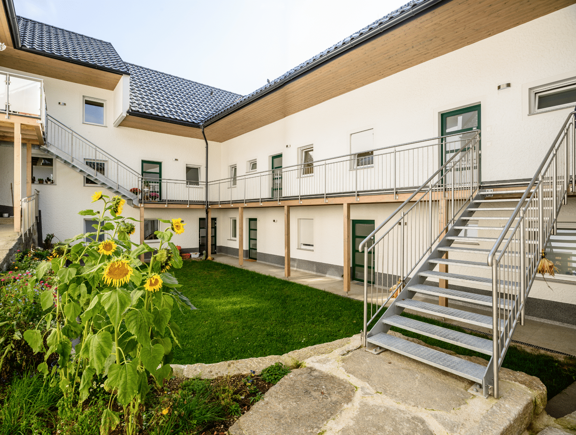 Bad Zeller zeigt den Innenhof eines Merhparteienhauses mit Edelstahl-Treppenaufgängen, Geländer, Sonnenblumen, grünen Fensterrahmen und Steinwegen.
