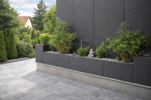 Eine Anordnung an rechteckigen Pflanzkübeln in Anthrazit mit passender Fassadenwand, bepflanzt mit Bambus, Zierpflanzen, hellem Kies mit kleiner Buddhafigur für ein abgerundetes Zengarten-Ambiente auf einer gepflasterten Terrasse in einem gepflegten Garten von Blumentrog-Flexi.