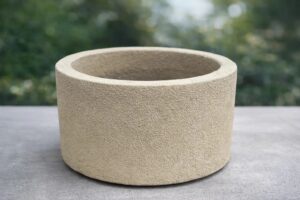 Ein moderner, runder Pflanzkübel in hellem Beige steht im Freien und ist schlicht, elegant sowie wetter- und UV-beständig.