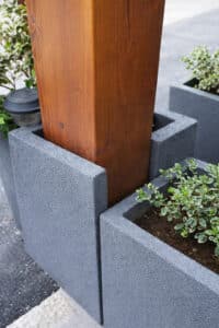 Blumentrog Flexi zeigt einen seitlich offenenen Pflanzkübel nach Maß in Anthrazit der sich an eine Holzäule schmiegt, angedacht zur Kletterbepflanzung.
