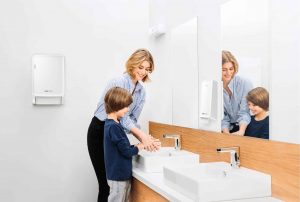 Frau hilft ihrem Kind beim Händewaschen in einem Bad mit CWS Pureline Produkten (Handtuschspender, Seifenspender, Aromadiffuser).