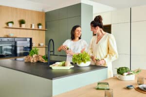 Zwei Freundinnen kochen in der Inselküche „Andros“ mit schwarzen Griffleisten, hängender Gerätezeile mit Holzdetails und größtenteils grifflose Küchenfronten in Grün, Olive oder Salbei, einer Kücheninsel mit schwarzer Arbeitsplatte und angedocktem Esstisch von DANKÜCHEN.