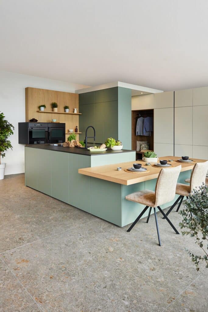 Inselküche „Andros“ mit schwarzen Griffleisten, hängender Gerätezeile mit Holzdetails und größtenteils grifflose Küchenfronten in Grün, Olive oder Salbei, einer Kücheninsel mit schwarzer Arbeitsplatte und angedocktem Esstisch von DANKÜCHEN.
