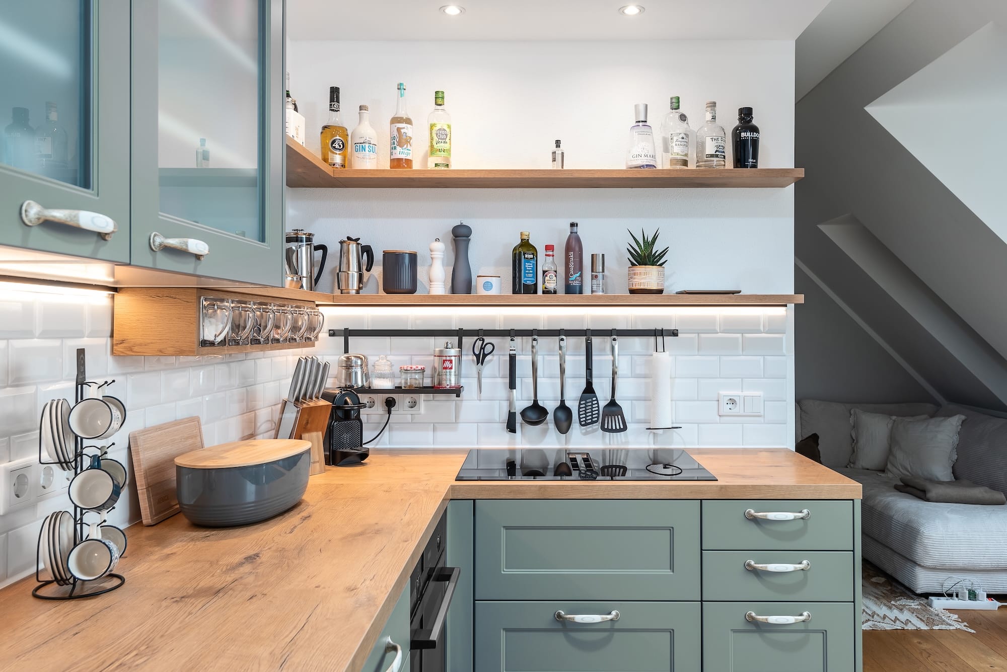 Landhausküche „Classico“ mit Kassettentüren in einem Grün wie Salbei oder Eukalyptus, Oberschränke mit Glaseinsätzen, antike silberne Griffe, Holzarbeitsplatte und gefliester Küchenrückwand mit Utensilienleiste von DANKÜCHEN.