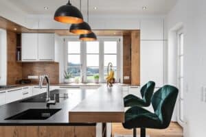 Eckküche „Living“ mit Insel, Spülbecken und Theke, teils grifflosen weißen Fronten, schwarzen Griffleisten, Holzoptik im Innenleben und Rückwänden, schwarzer Arbeitsfläche und Deckenspots von DANKÜCHEN.