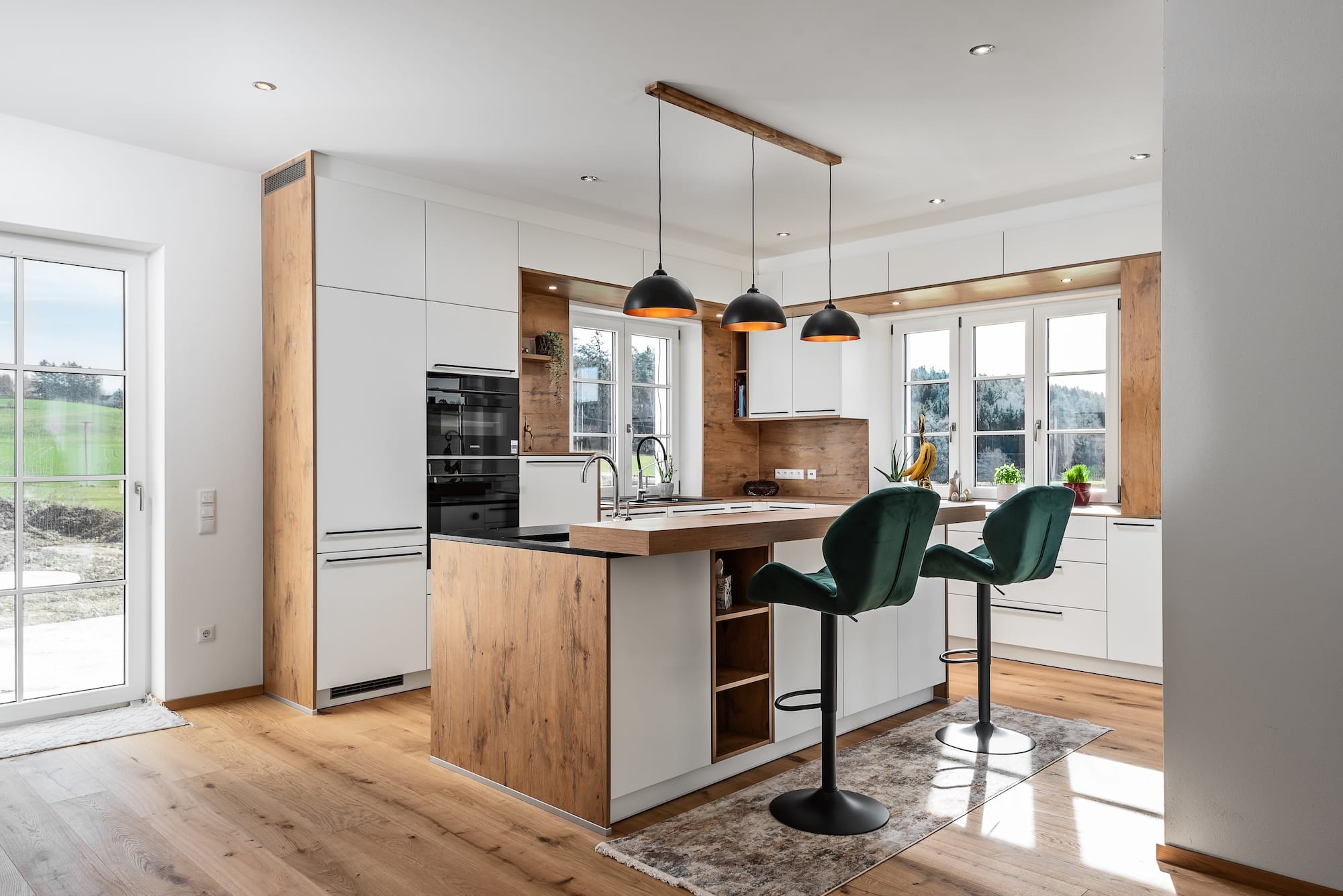 Eckküche „Living“ mit Insel, Spülbecken und Theke, teils grifflosen weißen Fronten, schwarzen Griffleisten, Holzoptik im Innenleben und Rückwänden, schwarzer Arbeitsfläche und Deckenspots von DANKÜCHEN.