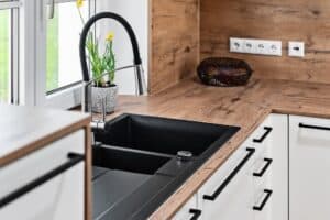 Eckküche „Living“ mit weissen Fronten, Spülbecken, Mischbatterie und Griffleisten in Schwarz und Arbeitsfläche und Rückwände in rustikaler Holzoptik von DANKÜCHEN.