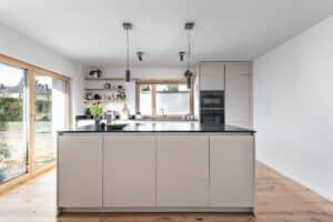 Moderne DAN-Inselküche Modell „Silbermond“ mit grifflosen Fronten in der Farbe Santa Cruz, schwarzer Arbeitsfläche, schwebenden Regalen, Holz-Akzenten und passenden Fensterrahmen.