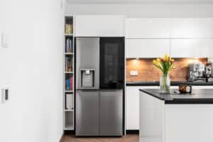 Inselküche „Silbermond“ mit grifflosen weißen Fronten, Gerätezeile mit Side-by-Side Kühlschrank, Holzoptik im Innenleben, schwarzer Arbeitsfläche und rustikalen Rückwänden von DANKÜCHEN.