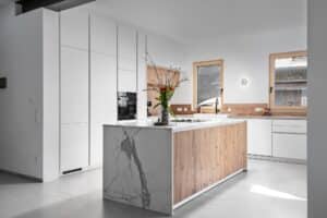 Moderne DAN-Inselküche Modell „Silbermond“ mit weißen grifflosen Fronten, rustikalen Holz-Akzenten, Kücheninsel, Seitenwand und Arbeitsplatte in Marmoroptik.