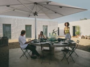 Innenhof eines mediterranen Apartmentkomplexes mit mehreren Bungalows und einer Aussenanlage mit Steinfundament, großem Steintisch, klappbaren Gartenstühlen und einem bodenmontierten, quadratischen Alu-Sonnenschirm in Weiss von DolenzGollner.