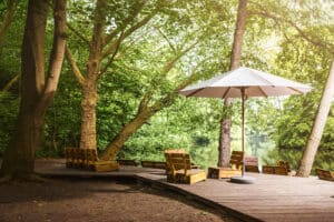 Waldlichtung mit hölzernem Terrassenpodest, niedrigen Sitzmöbeln aus Paletten gefertigt und einem freistehenden runden Holzsonnenschirm in Naturweiss mit Betonsockel von DolenzGollner.