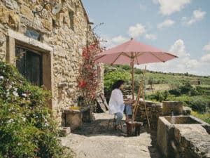Romantischer Eingangsbereich eines traditionellen, mediterranen Steinhauses mit einem freistehenden runden Holzsonnenschirm in einem Rotton der platzsparend eine Künstlerin und ihre Staffelei mit Gemälde vor Sonneneinstrahlung schützt von DolenzGollner.