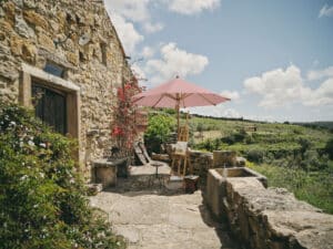 Romantischer Eingangsbereich eines traditionellen, mediterranen Steinhauses mit einem freistehenden runden Holzsonnenschirm in einem Rotton der platzsparend eine Staffelei mit Gemälde vor Sonneneinstrahlung schützt von DolenzGollner.