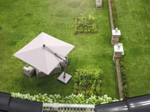 Gartengrundstück mit klassischer Ziegelmauer und einer Outdoorsitzreihe geschützt von einem freistehenden dunkelgrauen Sonnenschirm aus Aluminium mit Schwenkarm und leicht geneigt, aus der Vogelperspektive von DolenzGollner.