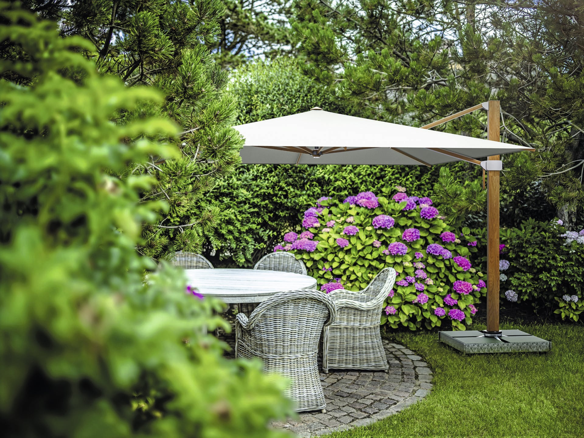 DolenzGollner Sonnenschutztechnik Ing. Wagner GmbH zeigt den Sonnenschirm "Aura" von Glatz in einem Garten mit blühenden Blumen und Rattan-Sitzmöbeln.