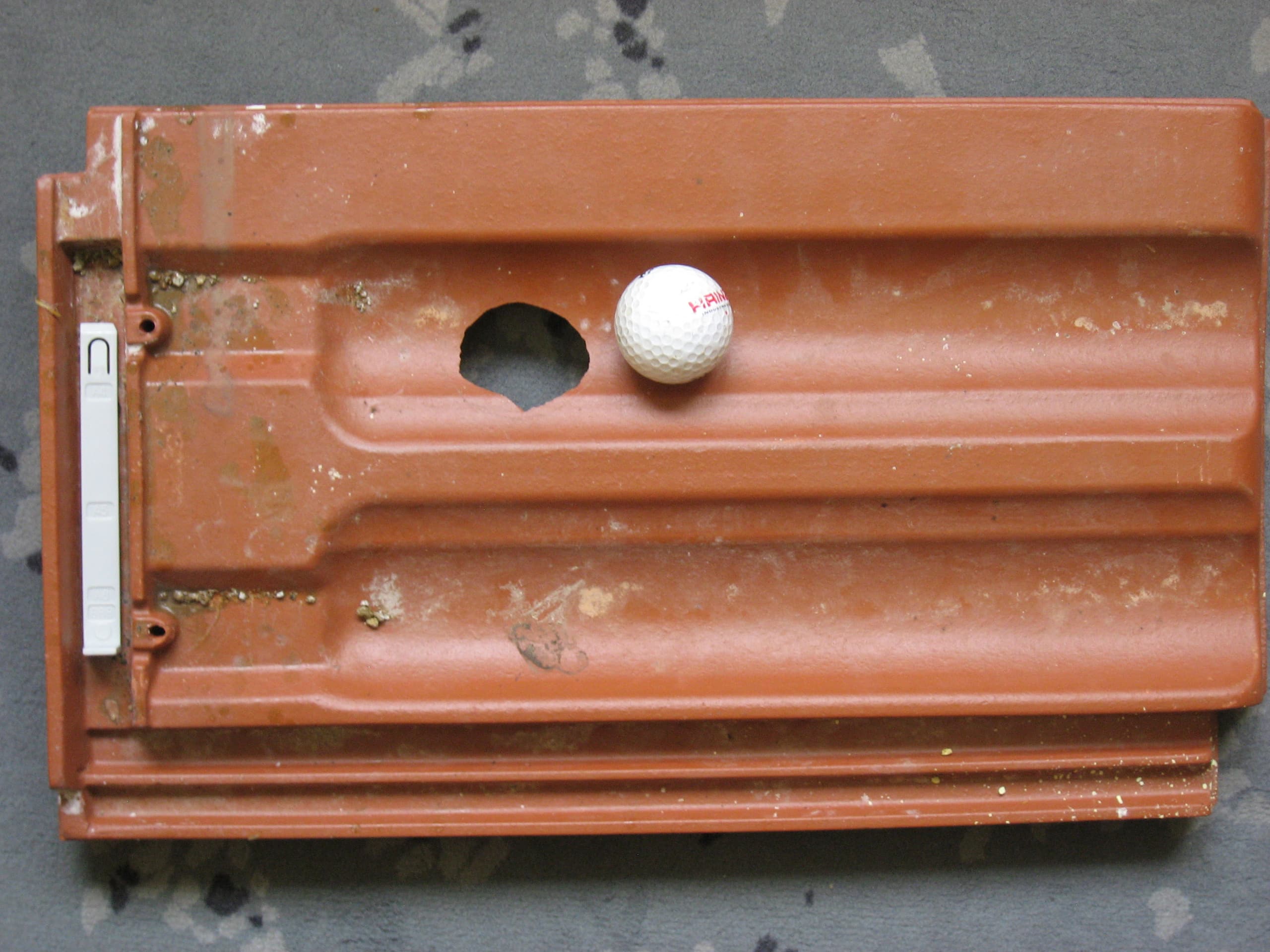Hagelschaden an einem Dachziegel mit einem Golfball als Größenvergleich bereitgestellt durch EPZ – Elementarschaden Präventionszentrum.