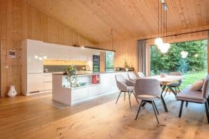 Erler Haus zeigt Wohnküche aus Küchenblock, Küchenzeile und angrenzendem Essbereich vor einer großen Glasfront in einem Raum mit Holzboden und Holwänden.