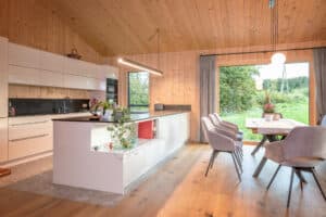 Offene Wohnküche mit Holzvertäfelung auf Decken und Wand, Küchenzeile und Kochinsel in Weiss, hochglanz, Esstisch mit Polsterstühlen und ein bodentiefes Alu-Holz-Fenster mit Ausblick in den Gartenvon ERLER Haus.