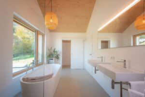 Badezimmer mit freistehender Badewanne, Doppelwaschbecken, Duschnische, einer holzverkleideten Decke und einem zweiflügeligen Fenster mit Holzrahmen von ERLER Haus.