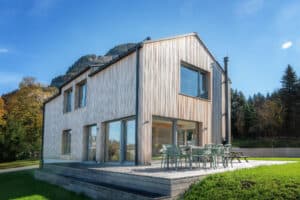 Einfamilienhaus mit Satteldach und vertikaler Holzsprossenverkleidung, mehreren Terrassenschiebetüren, bodenlose Fenster und Einfamilienhaus mit Satteldach und vertikaler Holzsprossenverkleidung, mehreren Terrassenschiebetüren, bodenlosen Fenstern und einem hölzernen Terrassenpodest mit Stufen von ERLER Haus. Terrassenpodest mit Stufen von ERLER Haus.