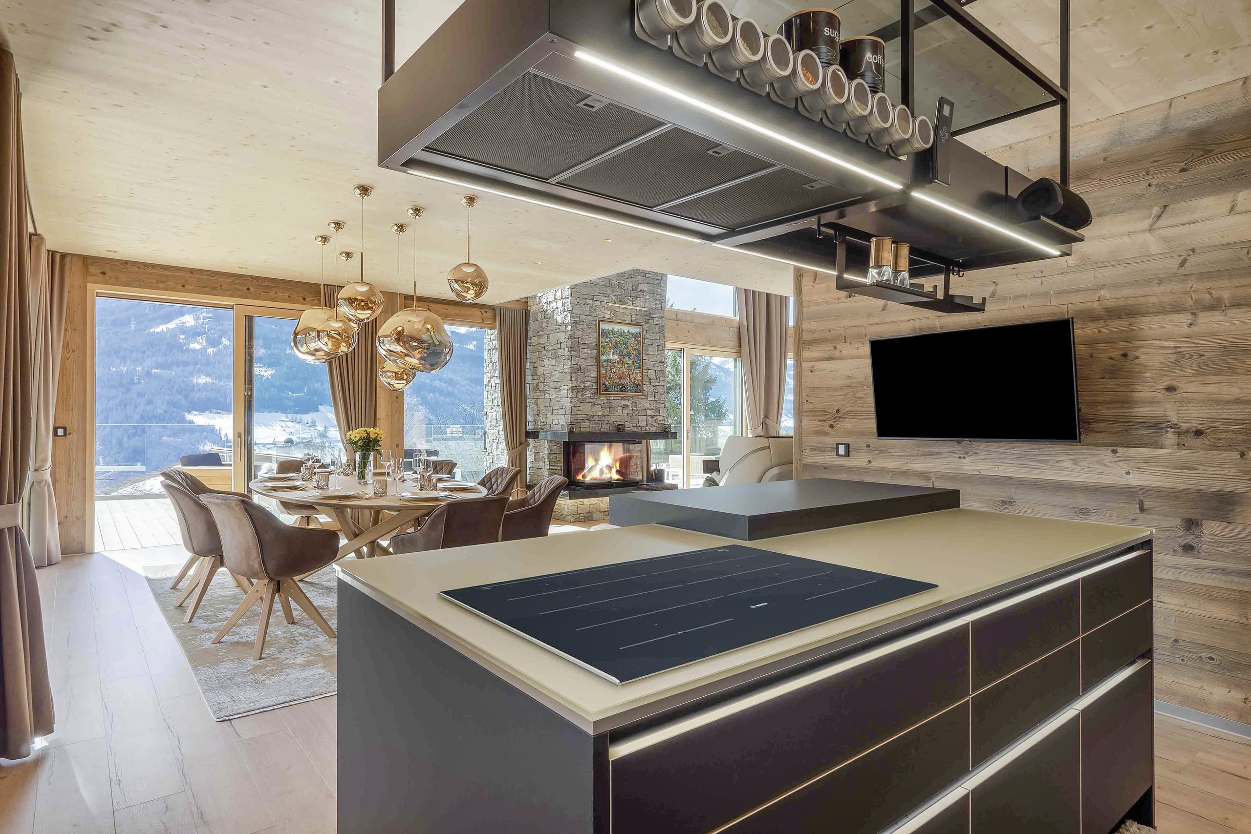 Offene Wohnküche eines Chalets mit Esstisch, Polsterstühlen, rustikalem Natursteinkamin und Terrassenschiebetüren und Verglasung mit Sicht in die Alpen von ERLER Haus.