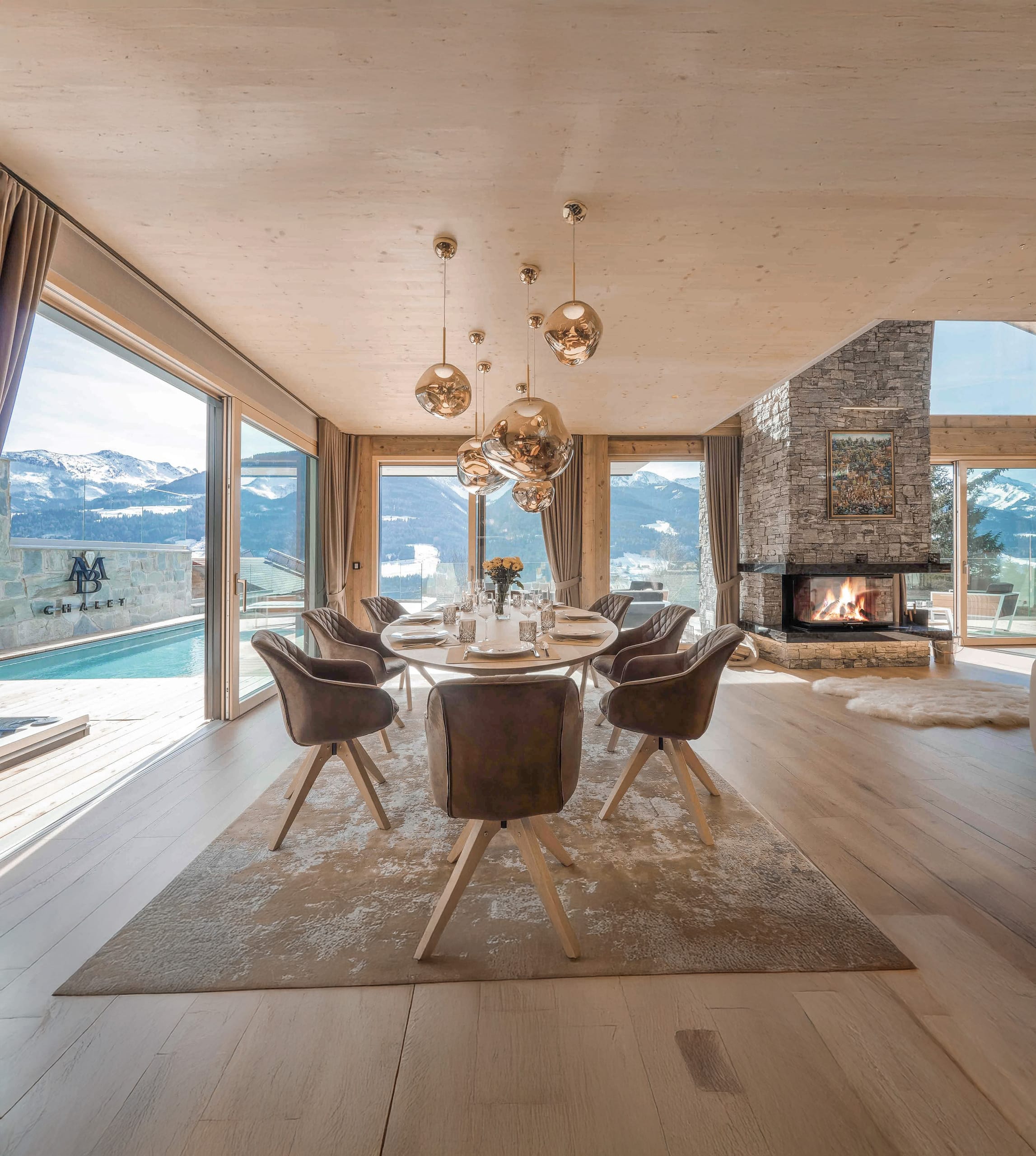 Offener Wohnbereich eines Chalets mit Esstisch, Polsterstühlen, rustikalem Natursteinkamin und Terrassenschiebetüren und Verglasung mit Sicht in die Alpen von ERLER Haus.