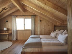 Ein gemütliches großes Schlafzimmer mit Holzdachschräge, Balken, Wandvertäfelung, Holz-Alu-Fenster, Doppelbett und einem wandmontiertem Holzschreibtisch von ERLER Haus.