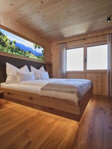 Ein komplett in Holz gehaltenes Schlafzimmer mit verkleideten Wänden, großem Bett mit Sockelbeleuchtung, Naturparkett und Holz-Alu-Fenstern von ERLER Haus.