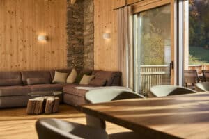 Ein offener Wohnraum im Holzhaus mit Esstisch und gepolsterten Stühlen, gemütlicher Couch, markantem Steinwandelement, Wandvertäfelung und passenden Holz-Alu-Terrassenschiebetüren von ERLER Haus.