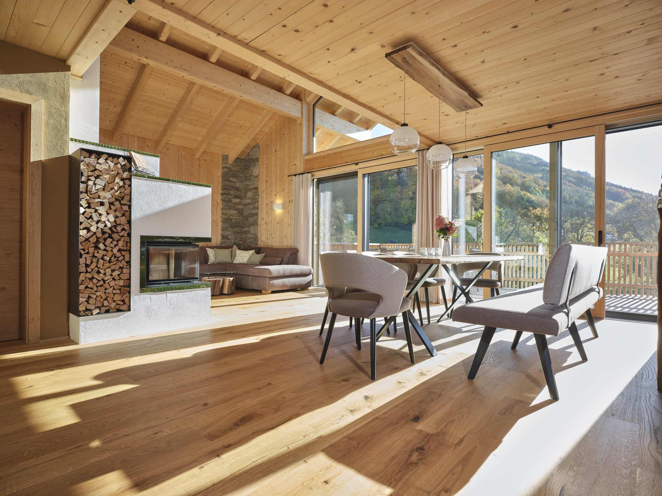 Ein offener Wohnraum mit Kamin und Holzlager, Esstisch mit gepolsterten Sesseln und Sitzbank, gemütlichem Sofa, Balkonschiebetüren und Holzdachstuhl in hochwertiger, natürliche Bauweise von ERLER Haus.
