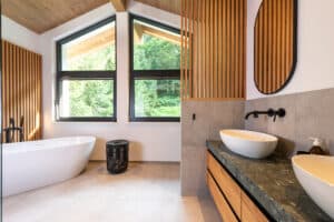 Ein modernes Badezimmer mit raumgetrenntem WC-Bereich, freistehender Badewanne, schwebendem Doppelwaschtisch, Holzsprossenelementen, Holzdecke, Dachschräge, großformatigen Fliesen und großen schwarzen Alu-Fenstern mit Oberlichten von ERLER Haus.